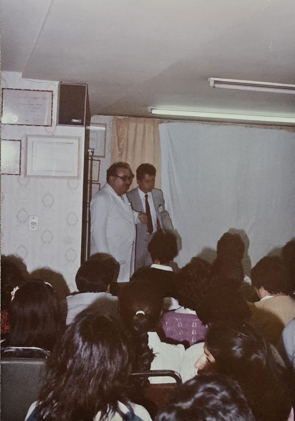 Profesor Dr. Francisco Arellano Ocampo en sesión académica enseñando dermatología en la Clínica Dermatológica de Puebla, presidiendo sala llena de jóvenes médicos en formación. Diplomas enmarcados visibles en pared al fondo.